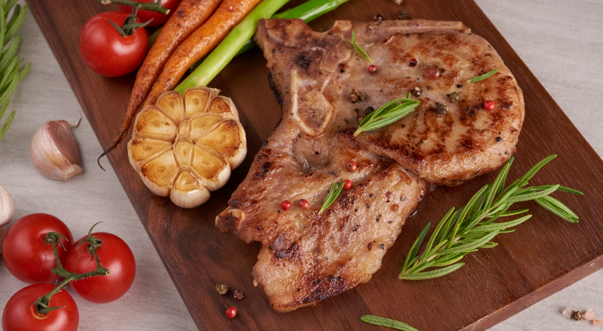 grilled-pork-steak-from-a-summer-bbq-served-with-vegetables-asparagus-baby-carrots-fresh-tomatoes-and-spices-grilled-steak-on-wooden-cutting-board-on-stone-surface-top-view.jpg